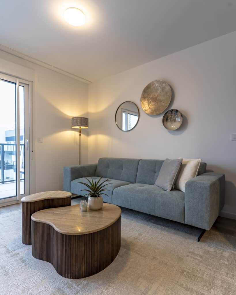 Moderne zithoek met blauwgroene bank, organische salontafels en wanddecoratie in natuurlijke tinten.