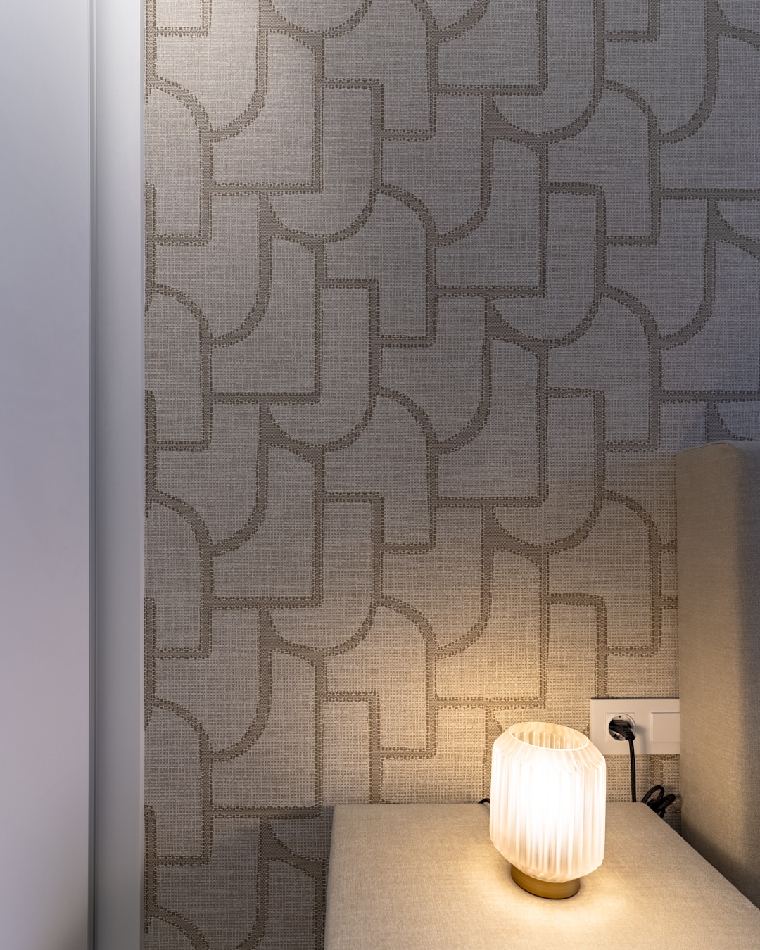 Nachtkastje met zachte stoffering, warme tafellamp en geometrisch behang als rustgevende combinatie in de slaapkamer