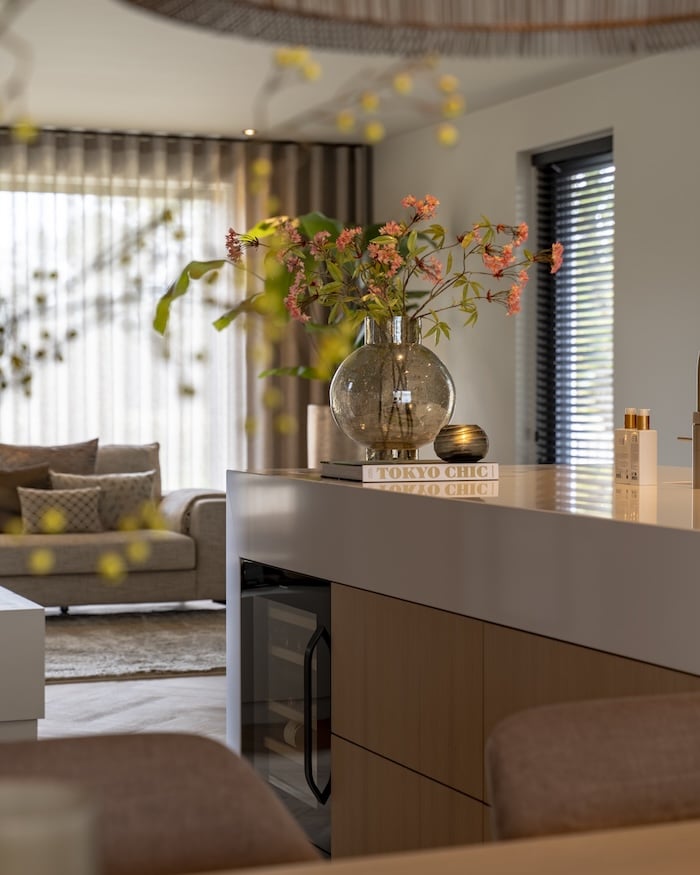 Ronde glazen vaas met roze bloemen op wit kookeiland, met zicht op zithoek