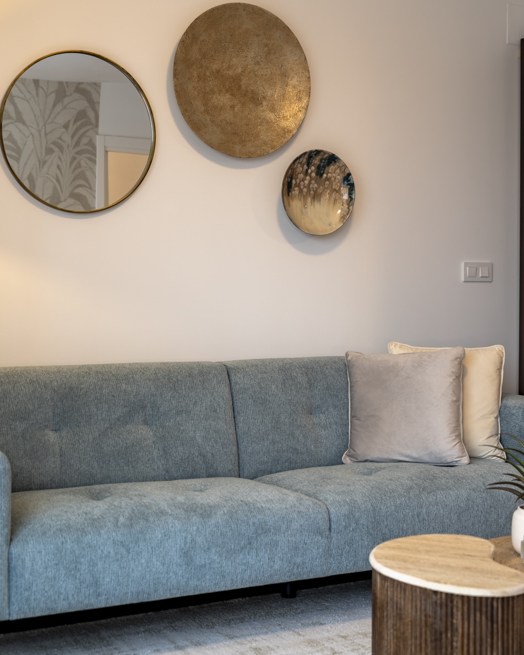 Gouden en bronzen wanddecoratie boven een lichtblauwe bank, met een glimp van het botanische behang via de spiegel.