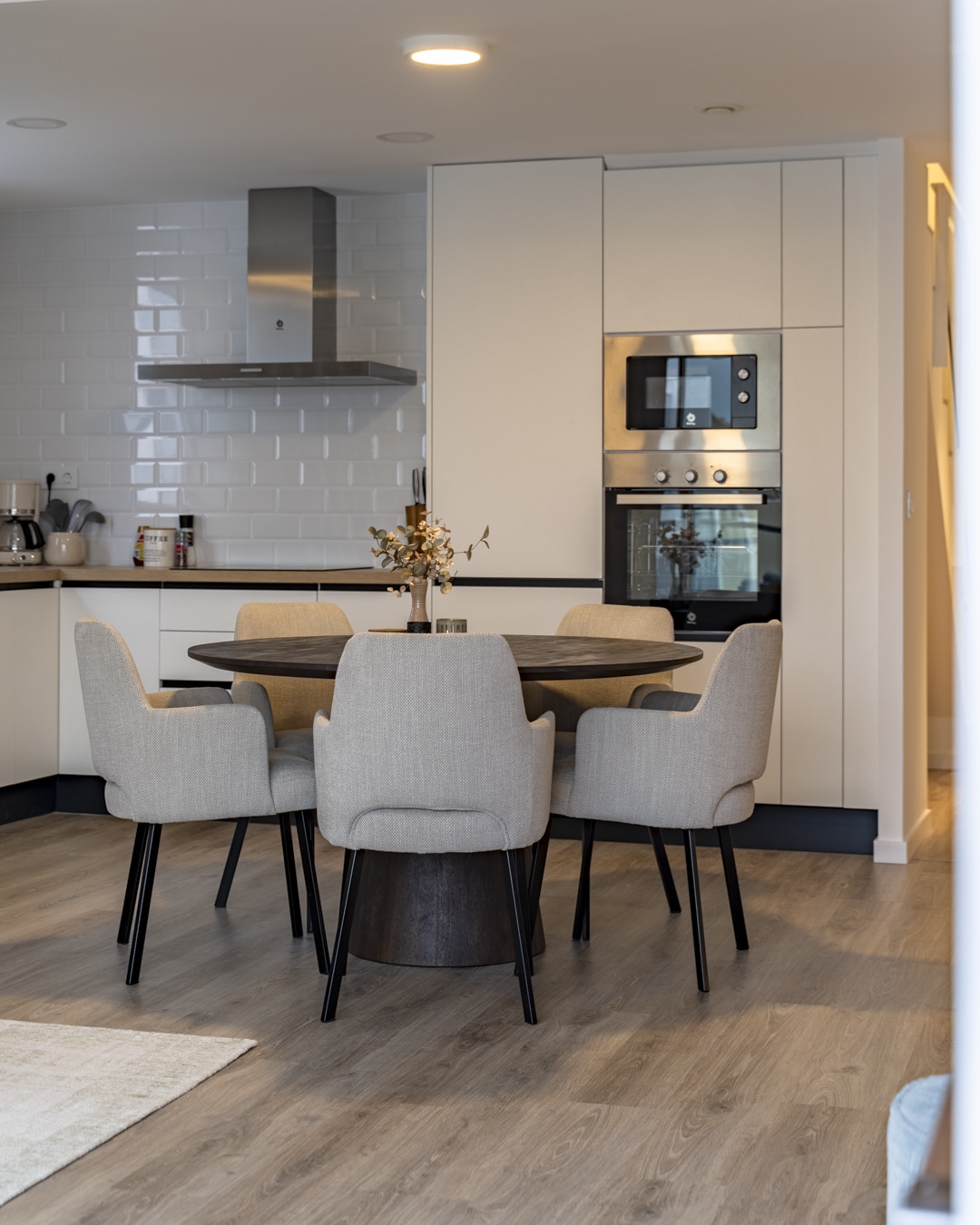 Open keuken met ronde eettafel en vijf gestoffeerde eetkamerstoelen op een lichte houtlook vloer.