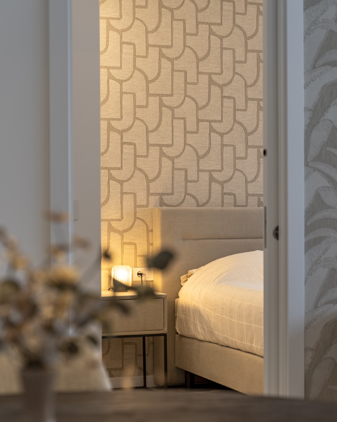 Zachte inkijk vanuit de woonkamer naar een slaapkamer met grafisch behang, warm licht en naturel tinten.