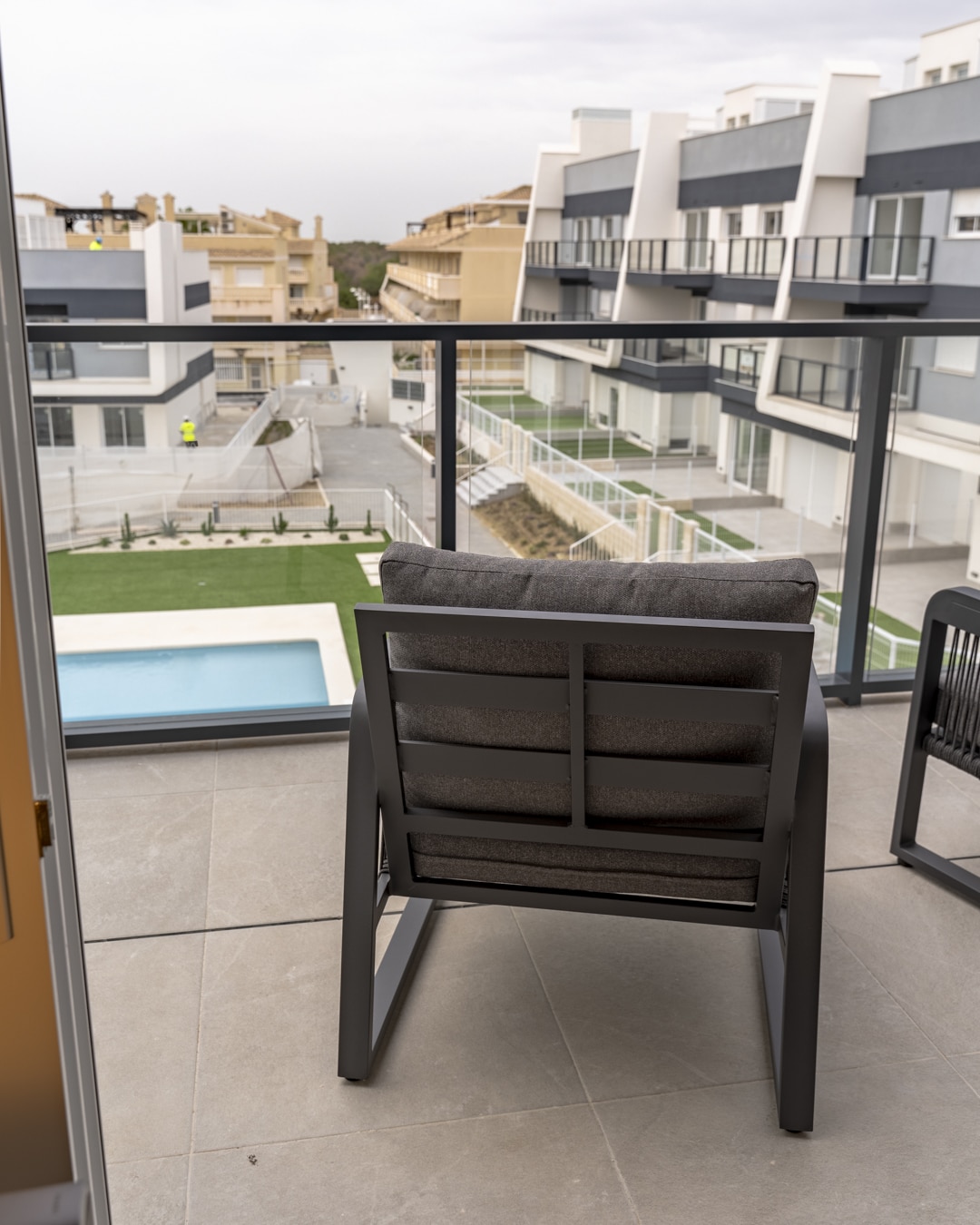 Zwarte loungestoel op balkon met zicht op zwembad en moderne appartementen in de verte.