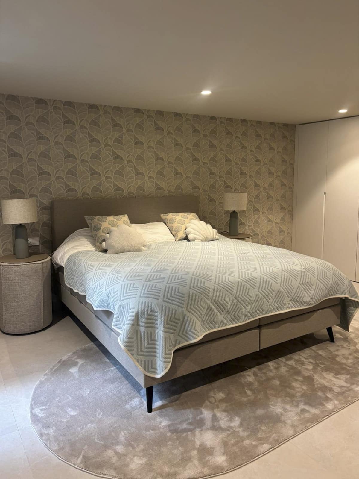 Stijlvolle slaapkamer met lichtblauwe bedsprei, luxe boxspring en modern dessinbehang in aardetinten, afgewerkt met bijpassende nachtkastjes en verlichting.