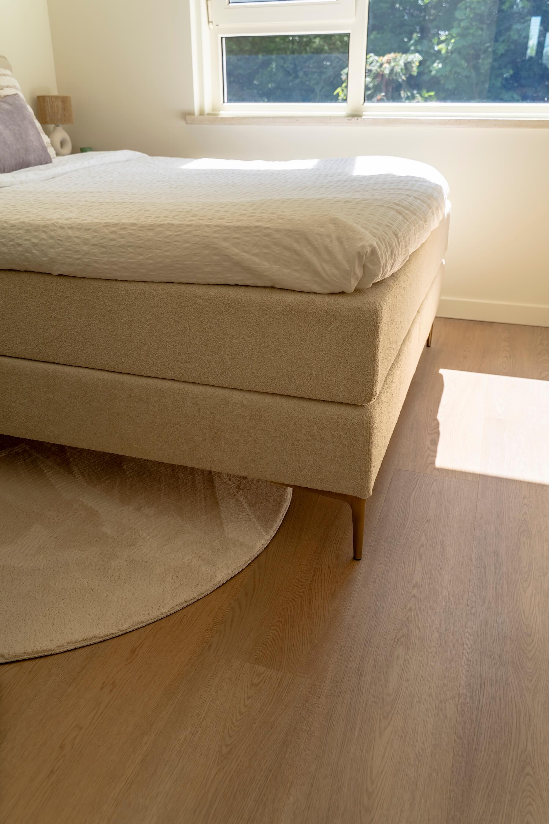 Onderkant van een boxspring en rond vloerkleed in een zonnige slaapkamerhoek