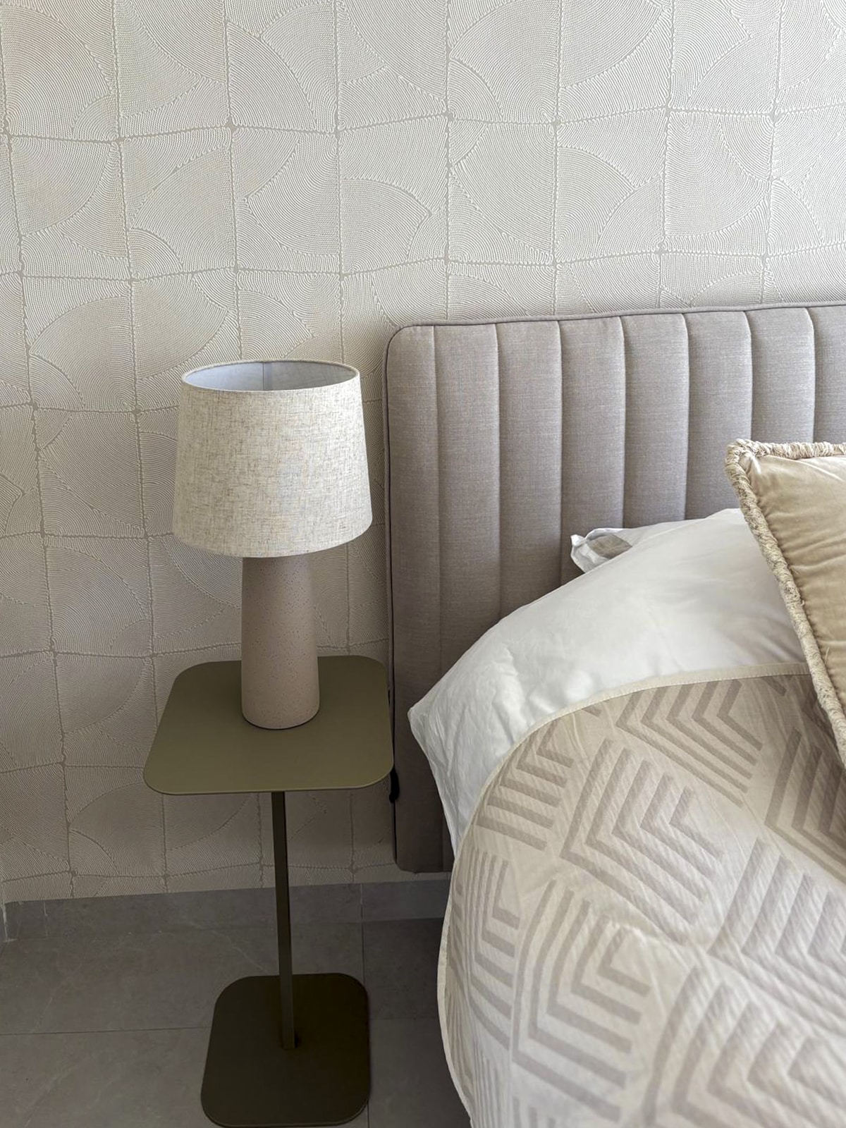 Close-up van nachtkastje en lamp met zachte materialen naast een gestoffeerd bed.