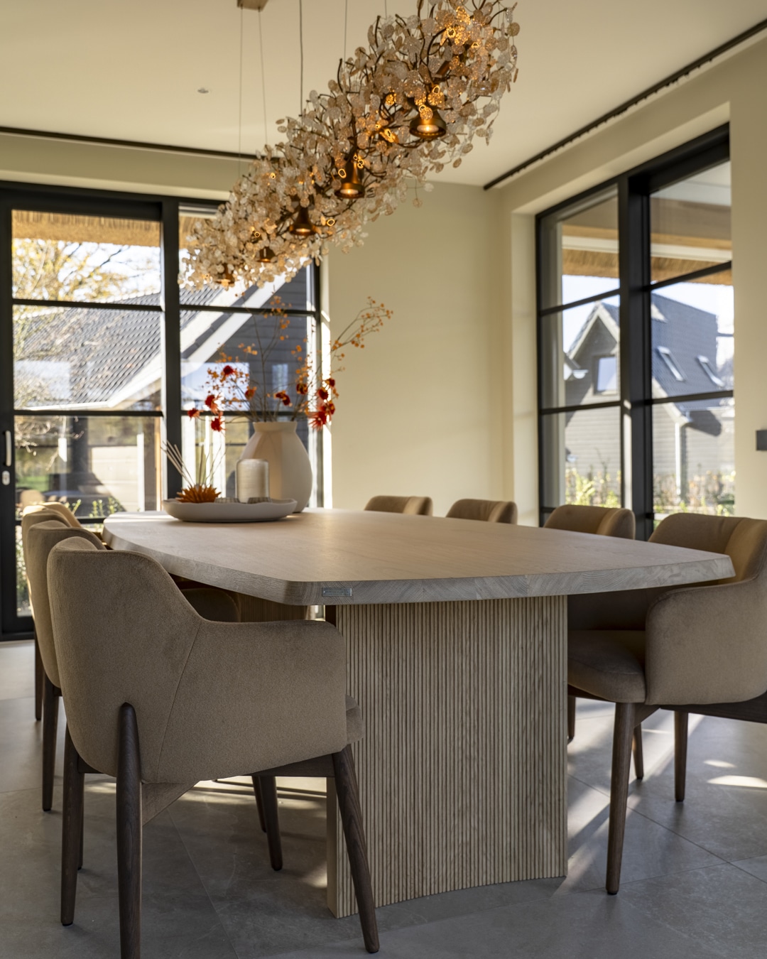 Detail van ovale eettafel en luxe gestoffeerde stoelen in natuurlijke tinten.